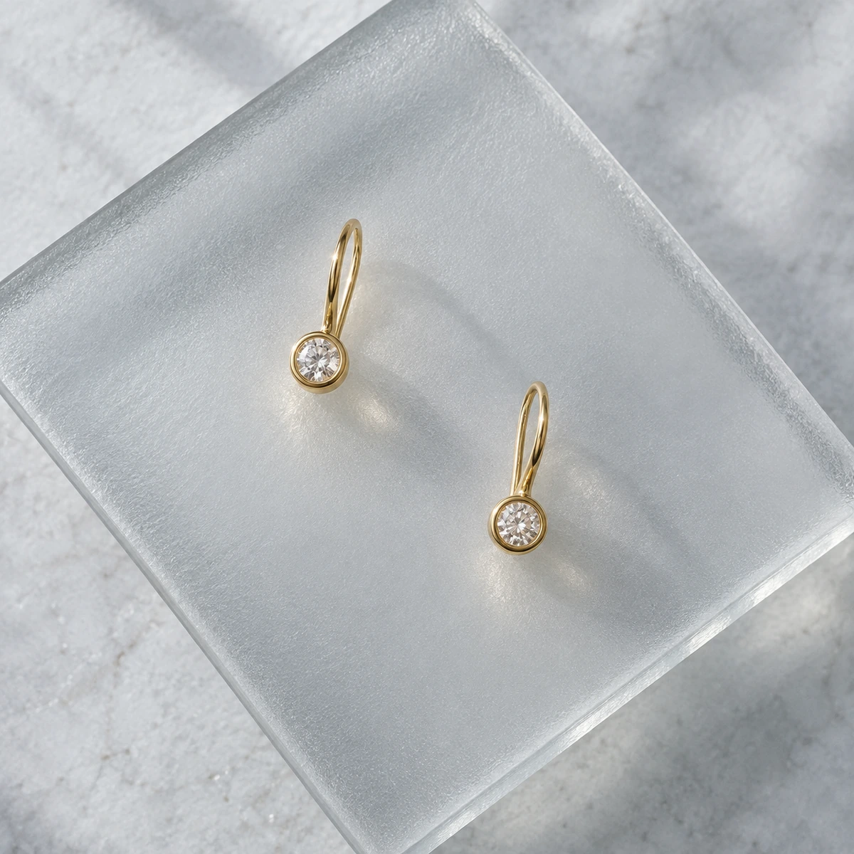Two gold earrings with small clear stones resting on frosted glass.